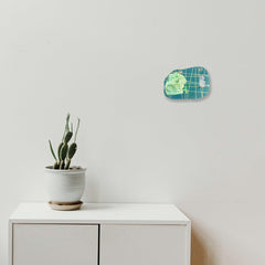 The artwork 'I Am Talking, Cat' by Sunny Chapman features a turquoise and lime green collage mounted on metallic paper with grid patterns, complemented by a modern minimalist setting showing a potted cactus on a white cabinet. Mixed media artwork combines monotype prints and marker details with grommet accents, mounted at 8x11 inches.
