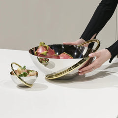 A person arranges the Anna New York Ring Fruit Bowl, a shiny stainless steel piece with gold accents filled with dragon fruit, as a centerpiece beside a matching smaller bowl holding strawberries on a white surface.