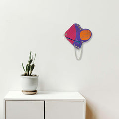 The artwork 'Satellite' by Sunny Chapman features an abstract geometric composition in vibrant magenta, purple, and orange hues on paper. The design incorporates curved shapes connected by a delicate silver chain, creating a modern, playful wall piece. The 12x12 inch acrylic painting contrasts boldly against a white background, by Sunny Chapman.