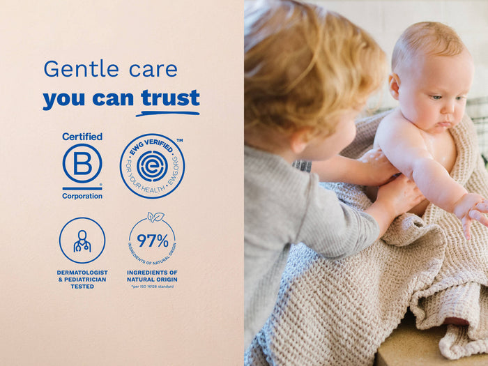 Mustela USA | Toddler applying lotion to a baby wrapped in a beige blanket.