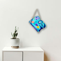 The artwork 'Coastal' by Sunny Chapman features vibrant abstract patterns in electric blue, neon pink, and bright yellow. Geometric shapes and fluid lines dance across the acrylic paper surface, creating a dynamic contemporary composition that evokes ocean waves and tropical energy. The piece is displayed on a white wall beside a potted cactus. By Sunny Chapman.