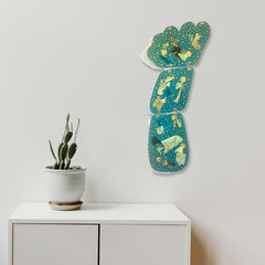 The artwork 'Dotter' by Sunny Chapman features a vertical series of connected turquoise panels with metallic gold speckles and abstract patterns, mounted on a white wall. Each panel showcases a unique collage design with shimmering metallic accents, created using monotype prints on premium paper stock, forming an elegant 40-inch vertical art piece.