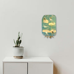 The artwork 'Flight' by Sunny Chapman features golden fish-like forms layered on turquoise striped metallic paper. The mixed media piece incorporates collaged monotype prints, suspended plastic charms, and metallic elements creating a whimsical aquatic composition displayed alongside a potted cactus on white furniture.