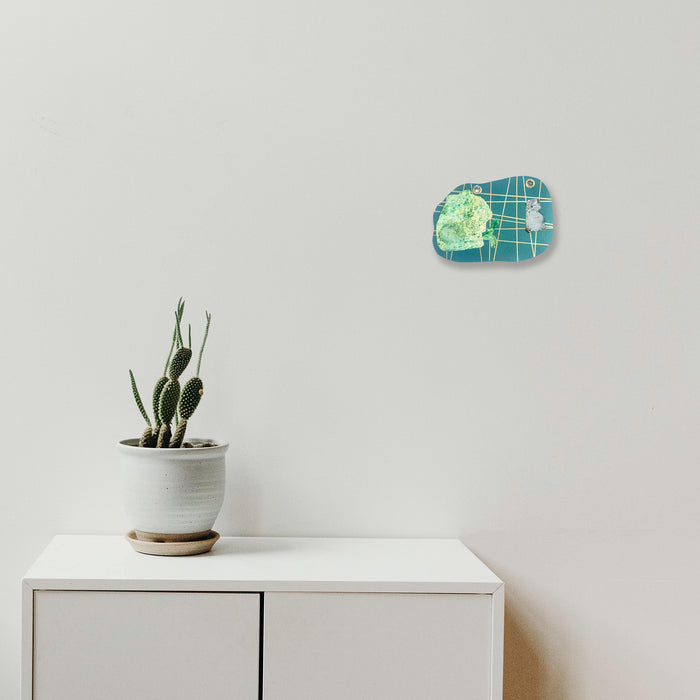The artwork 'I Am Talking, Cat' by Sunny Chapman features a turquoise and lime green collage mounted on metallic paper with grid patterns, complemented by a modern minimalist setting showing a potted cactus on a white cabinet. Mixed media artwork combines monotype prints and marker details with grommet accents, mounted at 8x11 inches.