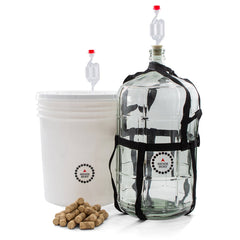 Master Vintner wine making fermenting setup with plastic bucket and glass carboy, both equipped with airlocks, plus a pile of wine corks in front.