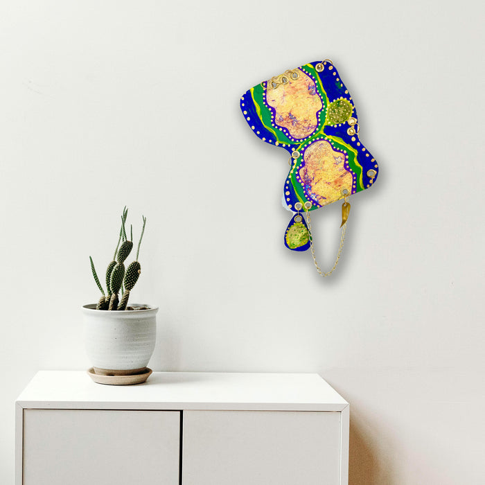The artwork 'Remembrance 1' by Sunny Chapman features an abstract organic shape in vibrant royal blue and emerald green borders, adorned with white dotted patterns. The central areas showcase peachy-gold iridescent sections, creating a luminous effect. The 12x16 inch acrylic painting appears to float against a white background, displayed above a modern cabinet with a potted cactus.