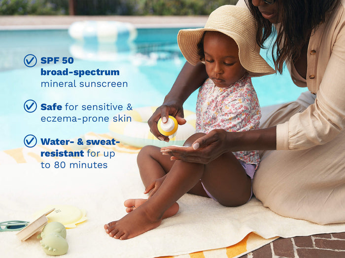 Mustela USA | Mother applying Mustela SPF 50 mineral sunscreen on her toddler's legs by a pool. Key benefits: broad-spectrum SPF 50, safe for sensitive and eczema-prone skin, water- and sweat-resistant up to 80 minutes.